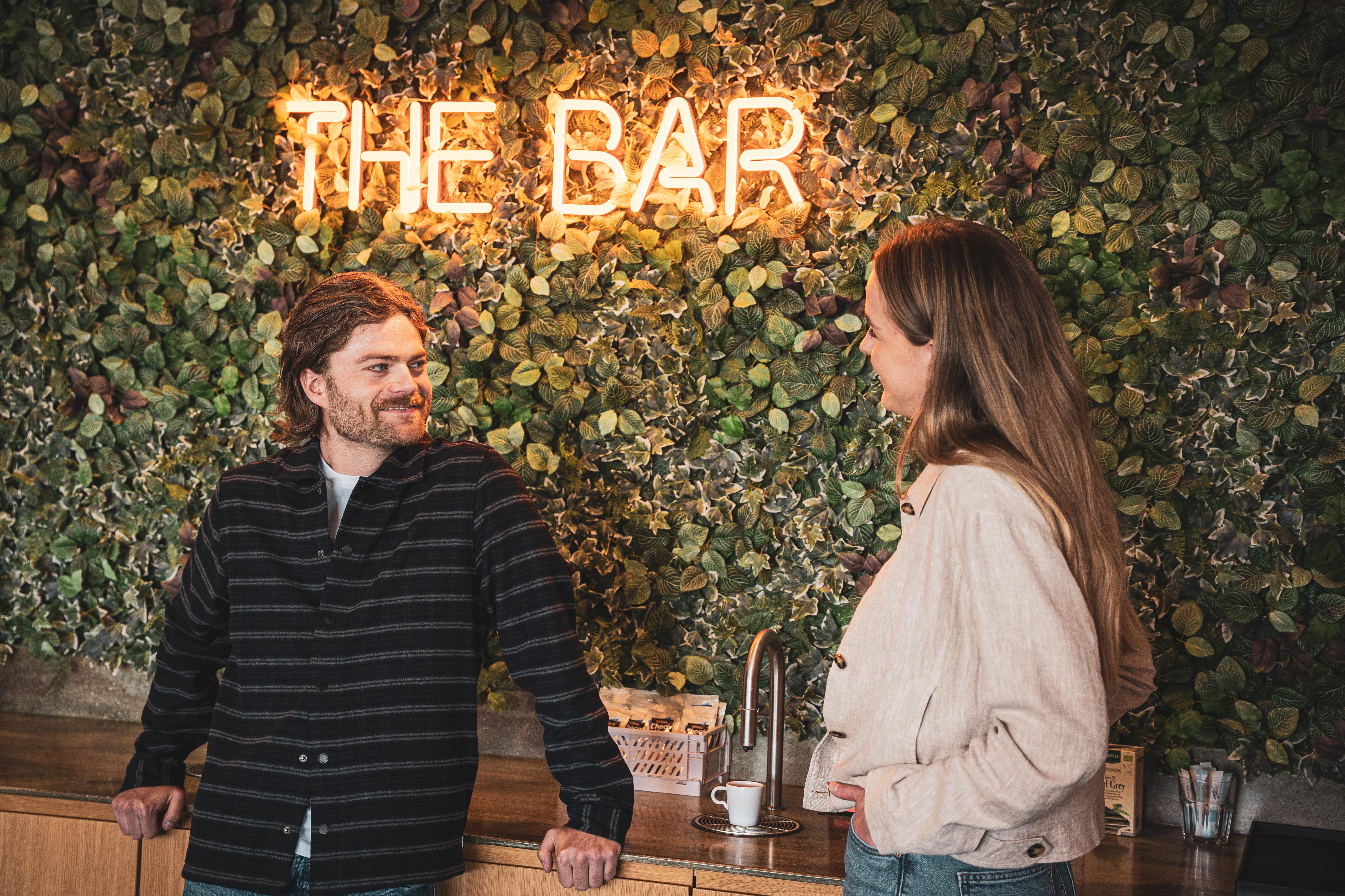 To unge taler sammen foran en grøn væg med neonskiltet 'THE BAR'