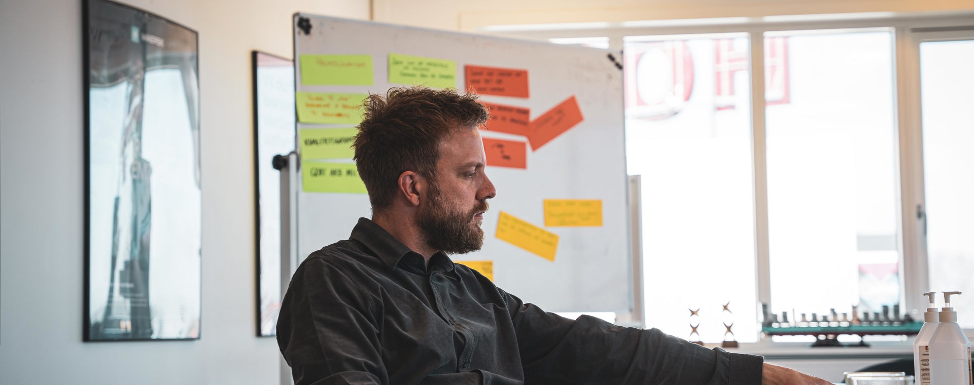 Underviser i sort skjorte ved bord med bøger og farverige post-it-sedler på whiteboard i baggrunden