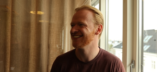Smilende mand i bordeauxrød t-shirt står ved vindue med gardin