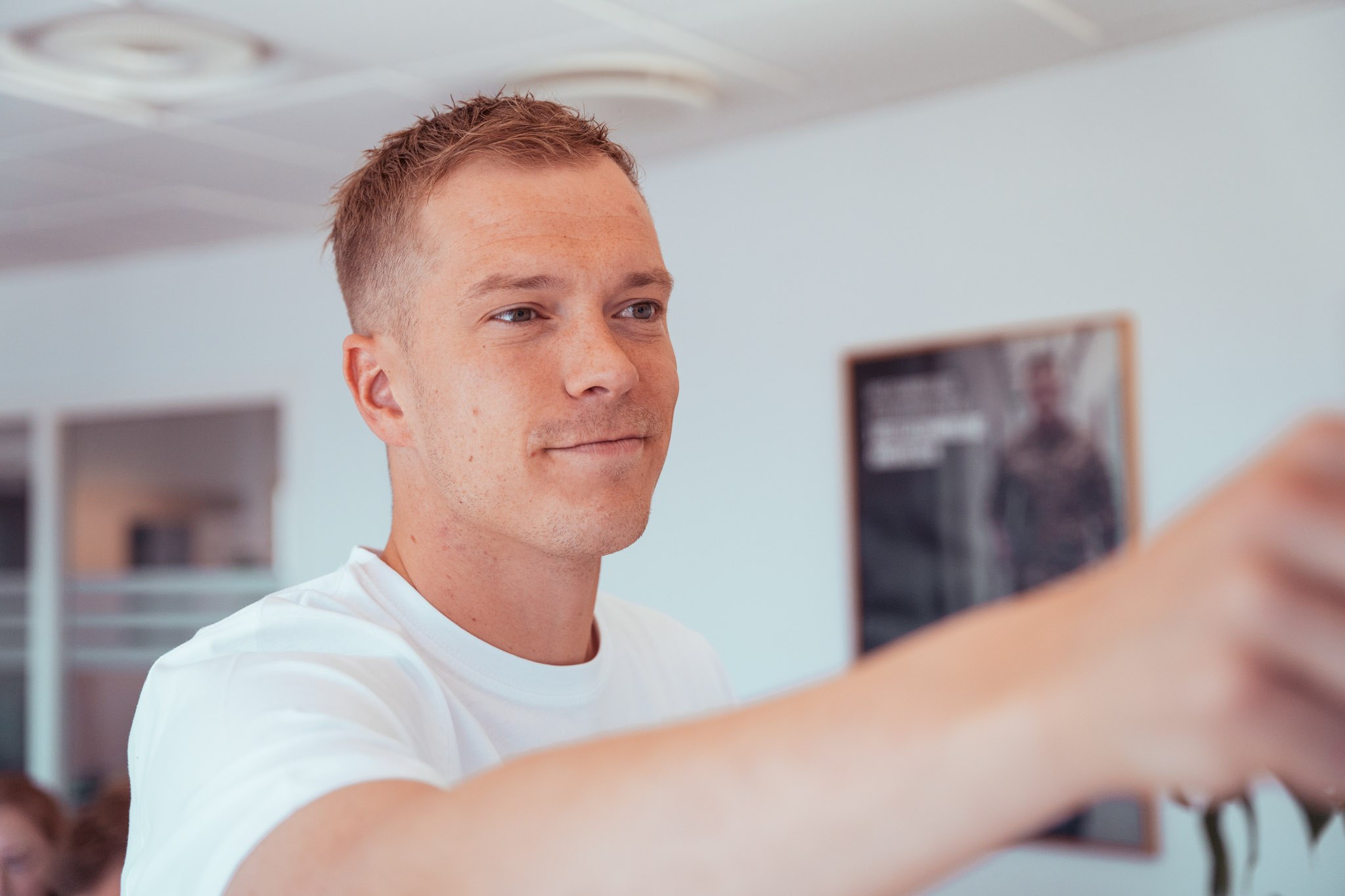 Ung mand i hvid t-shirt smiler og rækker armen frem i et lyst lokale