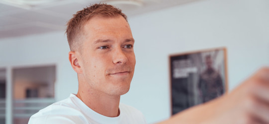 Ung mand i hvid t-shirt smiler og rækker armen frem i et lyst lokale